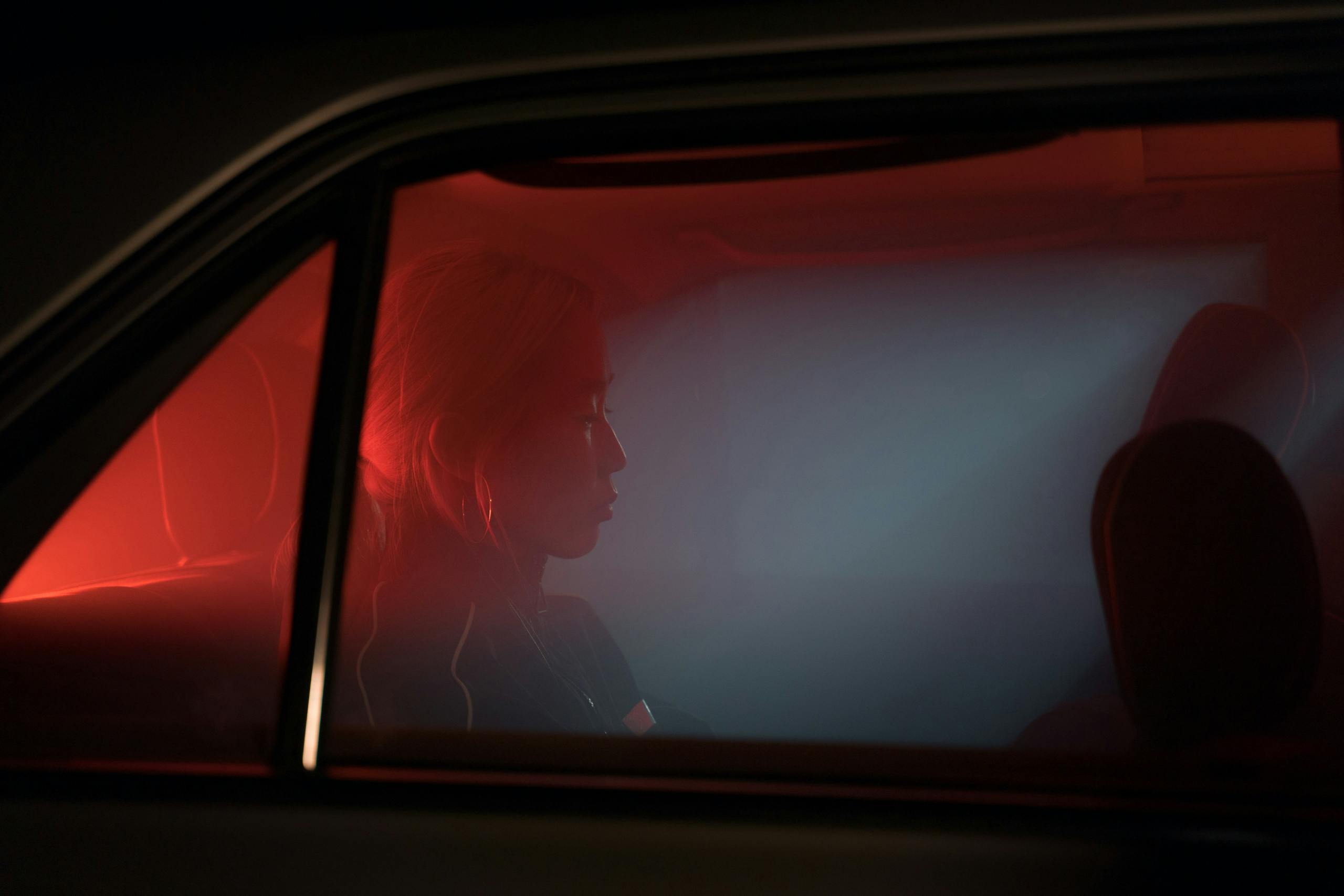 A woman sitting in a car, silhouetted against atmospheric red lighting at night, evoking solitude.